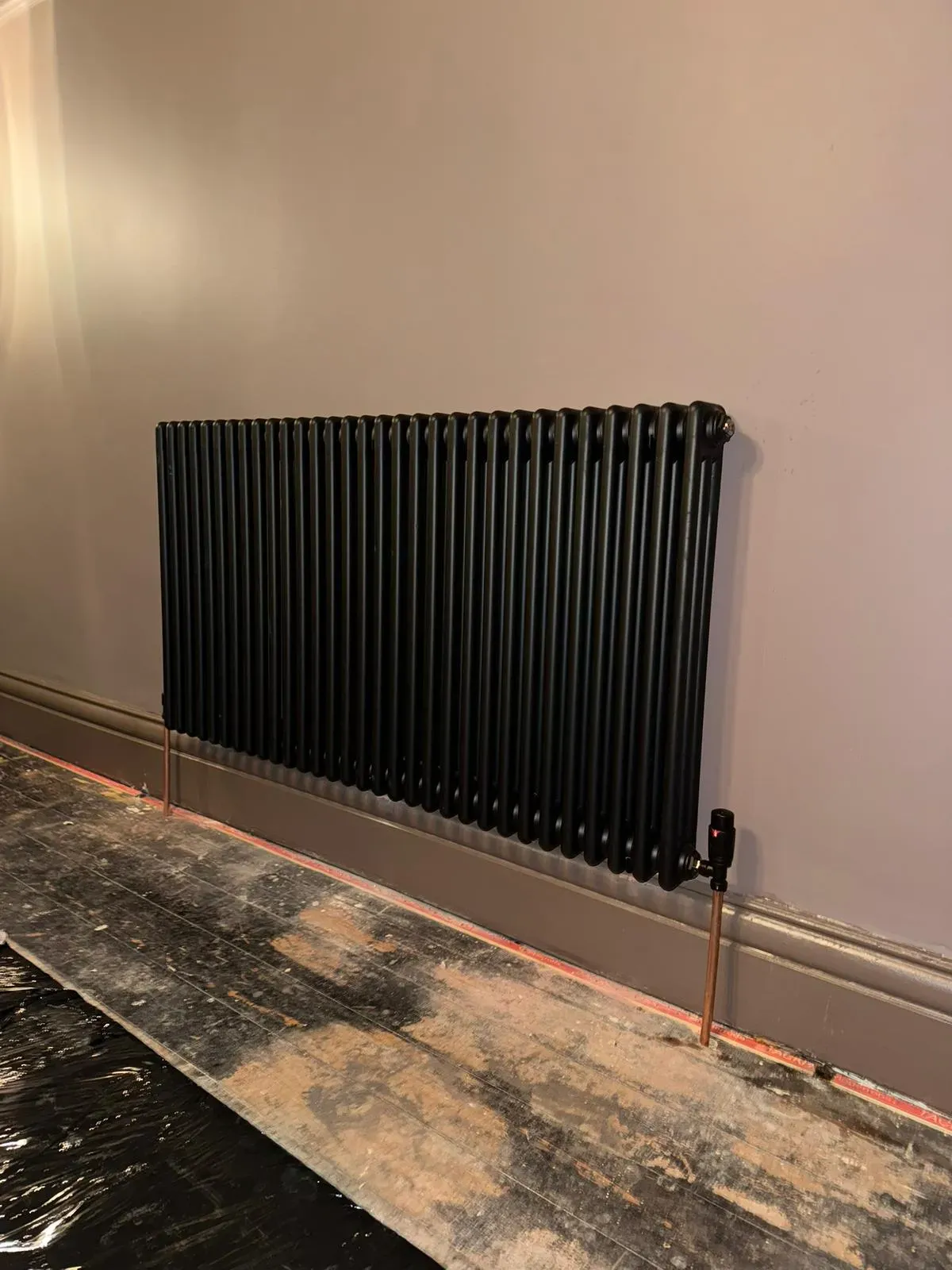 A modern black column radiator with copper pipework installed on a grey wall.